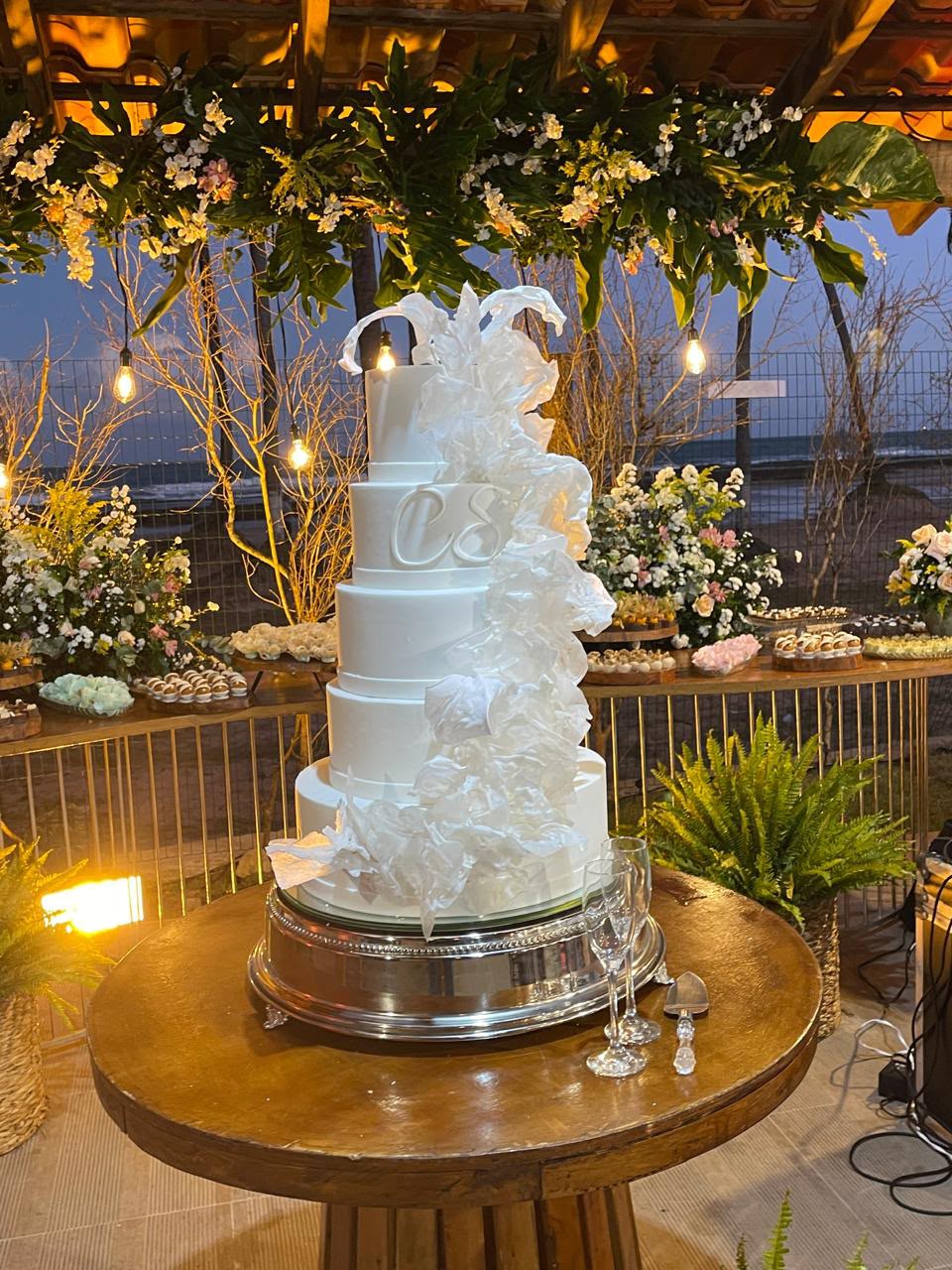bolo branco de vários andares com cascata de papel e monograma CS sobre mesa redonda de madeira, em decoração de festa à