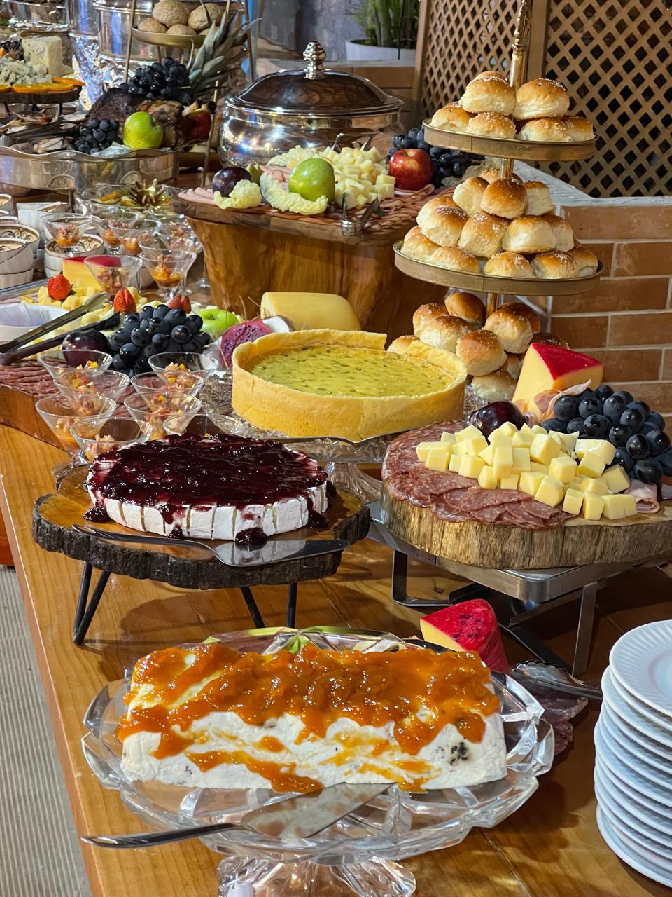 mesa farta de buffet com tortas, queijos, frios, frutas, pães em bandeja de três andares e taças de sobremesa