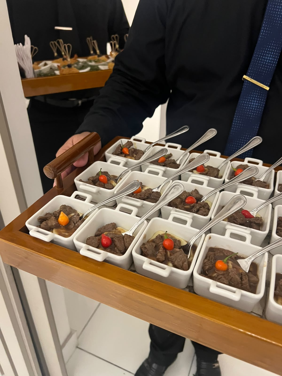 garçom de terno segurando bandeja de madeira com porções individuais de carne com pimenta e colheres em mini t