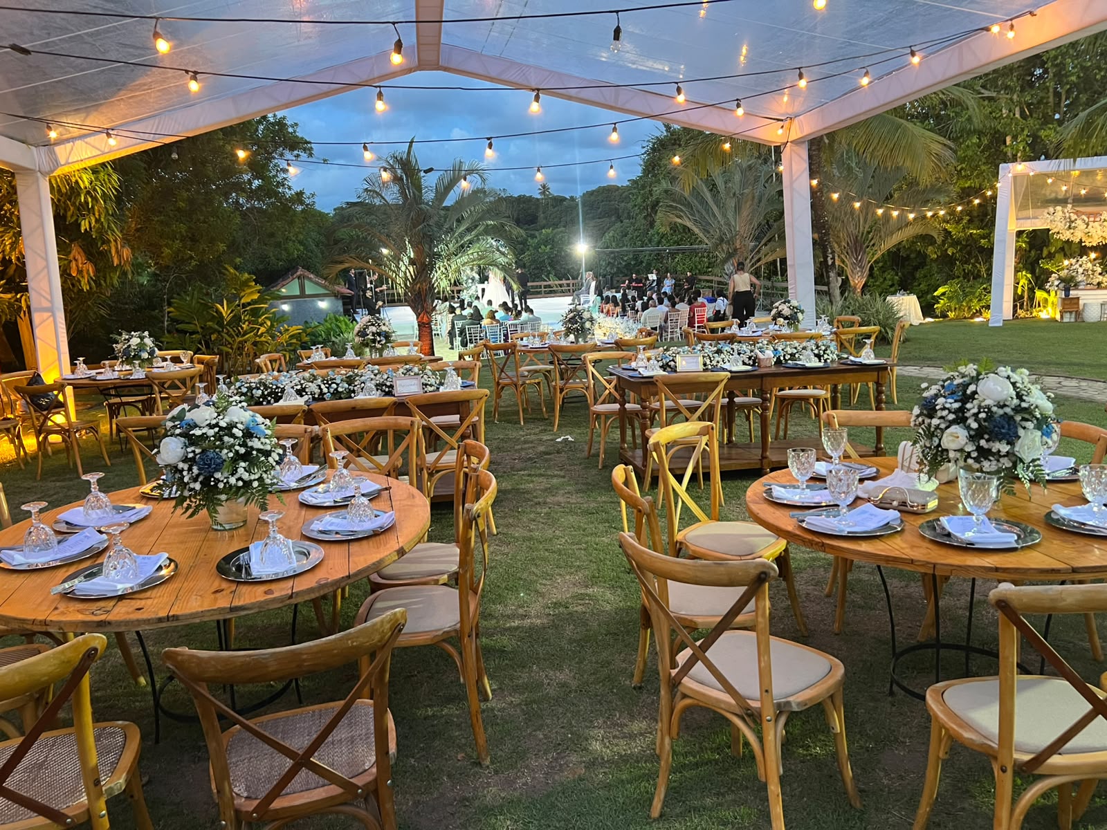 Decoração de evento ao ar livre sob tenda com luzes de fada, mesas redondas de madeira, cadeiras cross-back, a