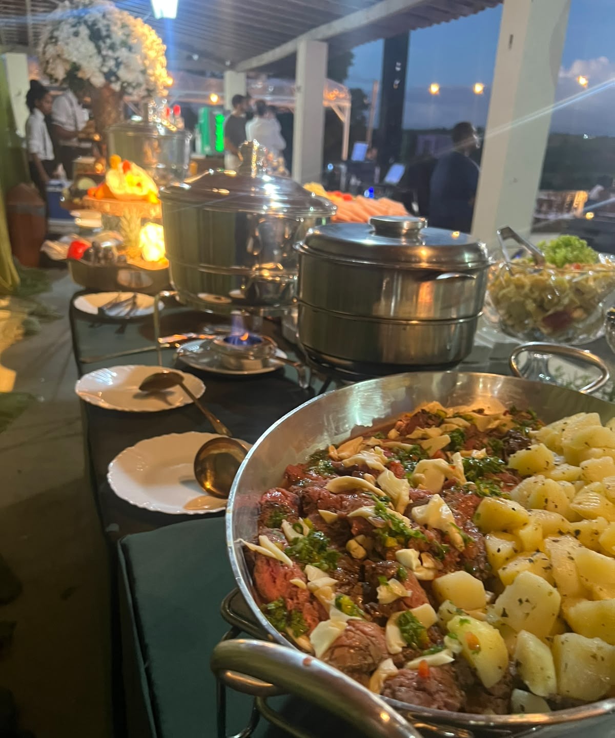 Mesa de bufê em evento ao ar livre ao entardecer com panelões de comida, frigideira grande com carne e batatas