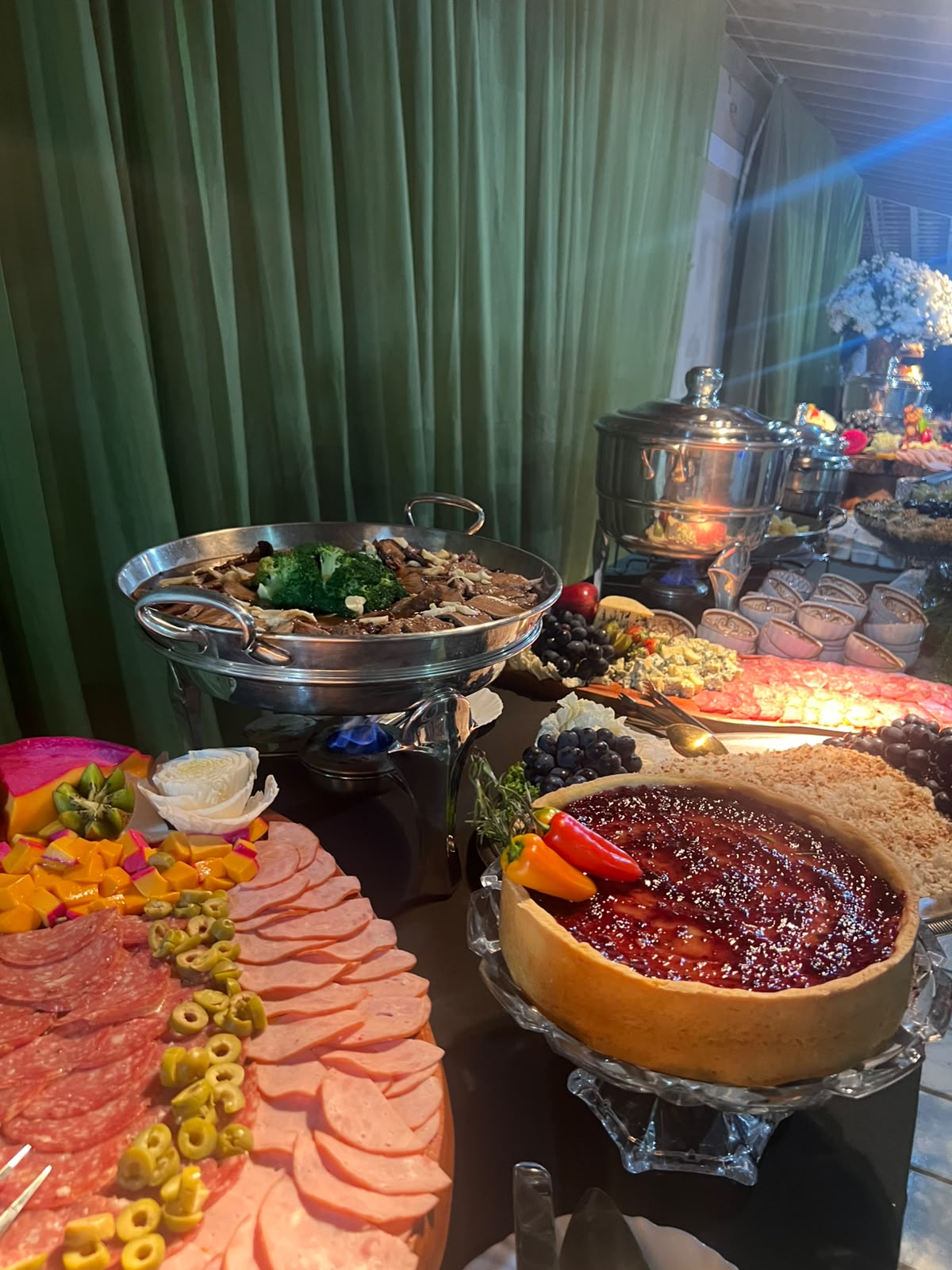 mesa de buffet de festa com tábua de frios, torta de geleia, queijos e rechaud com comida quente ao fundo de c